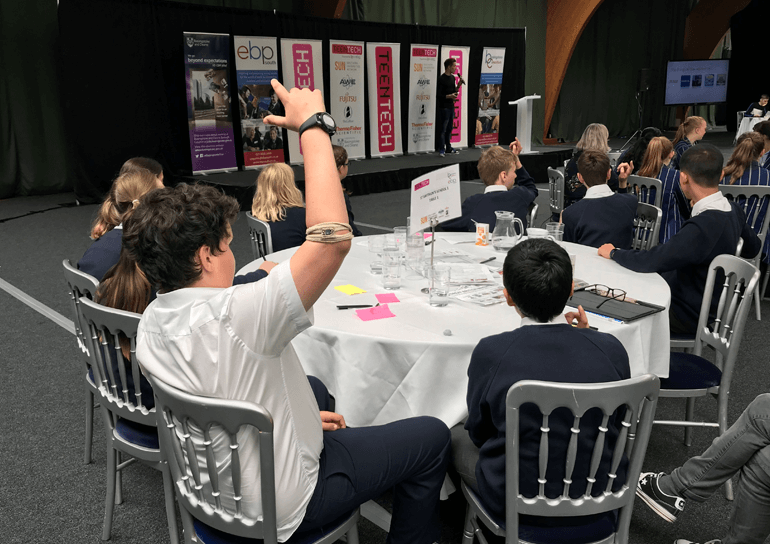 Leighton Park Boy in white shirt sat at table with hand up at TeenTech event