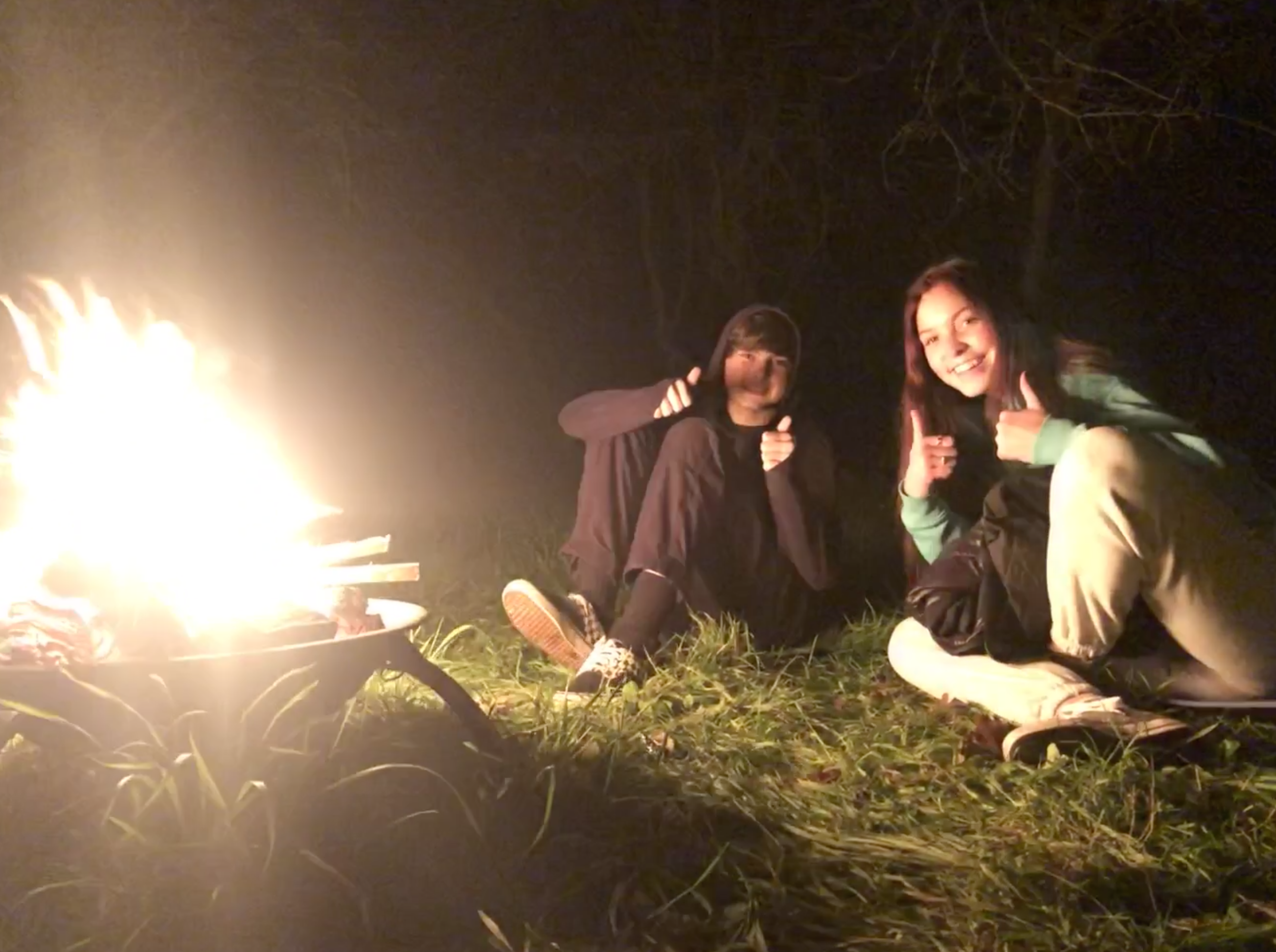 Two girls sat by a fire giving the thumbs up