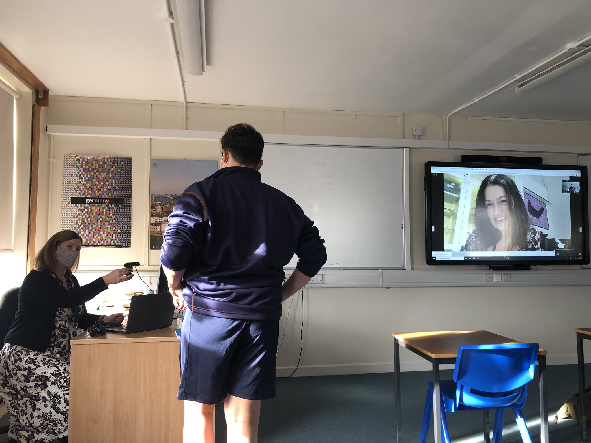 Student in blue jumper and shorts stood at a desk talking to a female teacher wearing a floral skirt