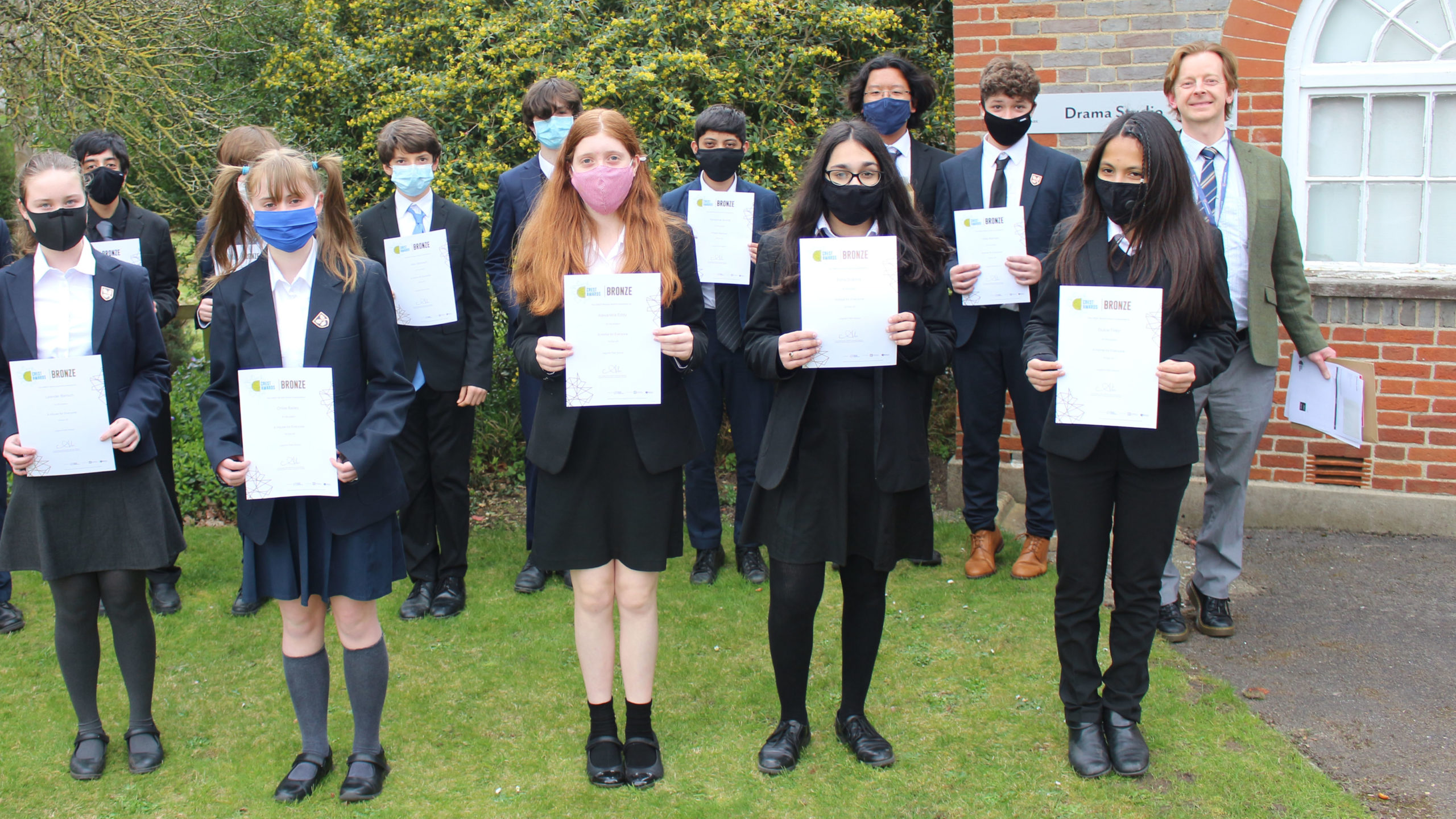 Students in uniform, wearing face masks stood holding certificates with teacher stood to the right of them